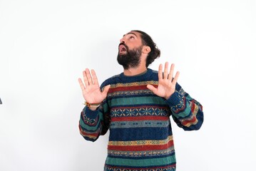 Fototapeta premium Caucasian man with beard wearing sweater over white background keeps palms forward and looks with fright above on ceiling tries to defense herself from invisible danger opens mouth.