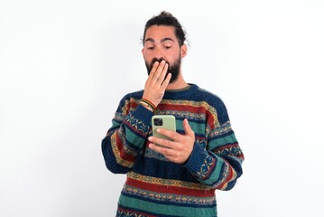 Caucasian man with beard wearing sweater over white background being deeply surprised, stares at smartphone display, reads shocking news on website, Omg, its horrible!