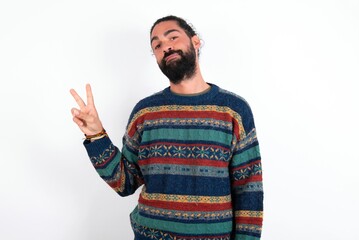 Naklejka premium Caucasian man with beard wearing sweater over white background makes peace gesture keeps lips folded shows v sign. Body language concept