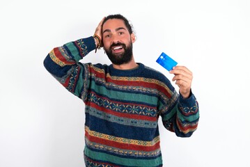 Impressed funny Caucasian man with beard wearing sweater over white background hand on head holding...