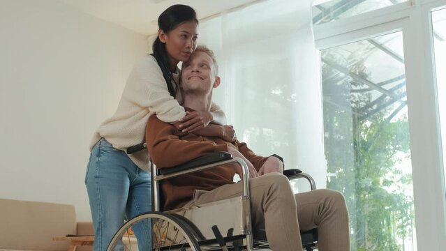 Young Caucasian wheelchaired man talking to his Asian girlfriend hugging him from behind