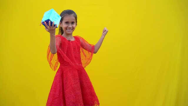 Portrait of a happy smiling girl showing a gift box isolated over yellow background. Smiling funny child in red dress holding Christmas gift in hand. Christmas concept.