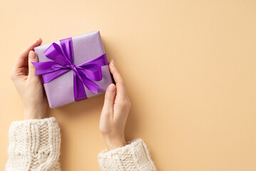 Christmas Day concept. First person top view photo of female hands in white knitted sweater holding lilac giftbox with violet ribbon bow on isolated pastel beige background with copyspace
