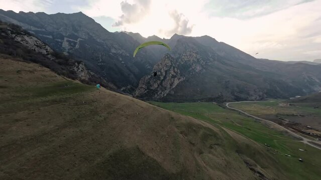 FPV drone follow to man paraglider flying extreme sport activity at mountain rocky terrain valley enjoy freedom. Aerial view shot paraplane parachute outdoor adventure over cliff summit sky scenery 4k