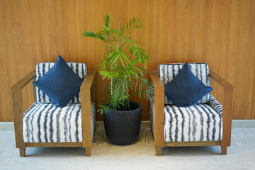 Twin wooden chairs with foam and pillows in a hotel lobby decorated with green plants in pots.