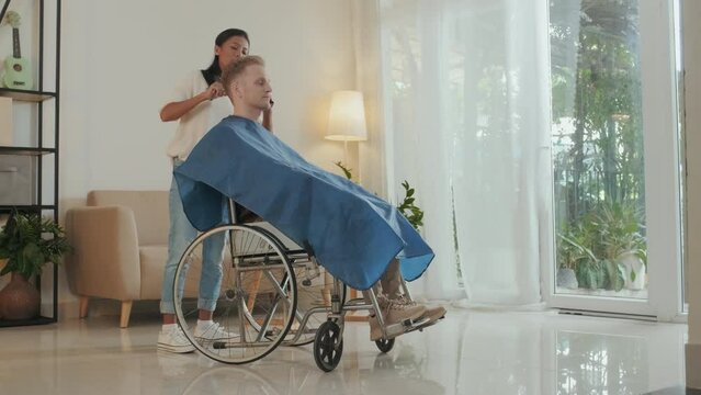 Caucasian wheelchaired man having his hair done by his Asian girlfriend at home