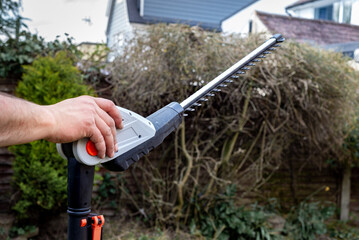 Close up of battery powered hedge clippers with man's hand holding it in front of hedge in garden.