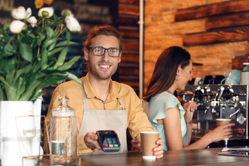 Young business owner with cup of coffee and payment terminal in his cafe