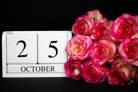 October 25 wooden calendar, white on a black background, pink roses lie nearby.Postcard with copy space. The concept of a holiday, congratulation, invitation, party, announcement, vacation,promotion.