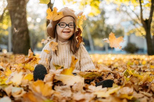 Happy cute brown-haired woman with Down's syndrome in a knitted sweater and stylish glasses throws bright foliage into the sky and laughs, the child enjoys time in a warm autumn park, happy childhood
