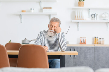 Naklejka na ściany i meble Thoughtful mature man with pen and clipboard sitting at table in kitchen