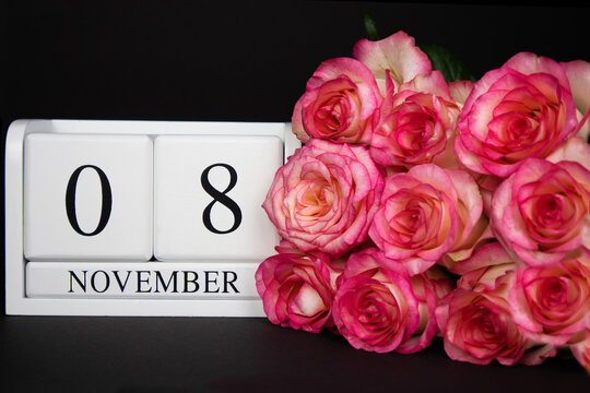 November 8 wooden calendar, white on a black background, pink roses lie nearby. Postcard with copy space. The concept of a holiday, congratulation, invitation, party, announcement, vacation,promotion