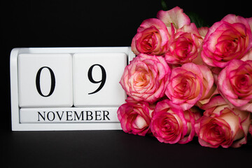 November 9 wooden calendar, white on a black background, pink roses lie nearby. Postcard with copy space. The concept of a holiday, congratulation, invitation, party, announcement, vacation,promotion.