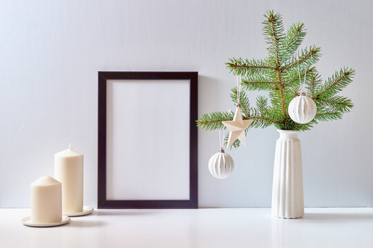 Mock up black poster frame with christmas decoration in home interior, scandinavian style. Green fir branches in a vase and ball on a white table
