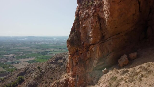 via ferrata. Man climbing mountain, drone view, traveling in to general shot of the climber, we see an orange mountain and some crop fields in the background