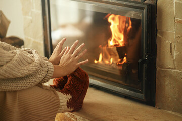 Woman in cozy wool socks warming up feet and hands at fireplace in rustic room. Heating house in...