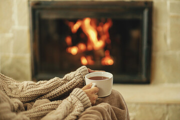 Hands in cozy sweater holding cup of warm tea on background of burning fireplace close up, autumn...