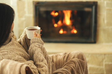 Fototapeta na wymiar Woman in cozy sweater holding cup of warm tea at fireplace in rustic room. Heating house in winter with wood burning stove. Young stylish female relaxing at fireplace in farmhouse