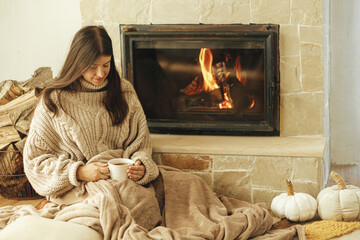 Woman with cup of warm tea relaxing at fireplace, autumn hygge. Heating house with wood burning...