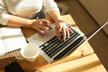 Fototapeta na wymiar Cropped shot top view of female hands typing on keyboard of opened laptop at kitchen table with white coffee mug and notebook, studying online, checking e-mail, using learning application