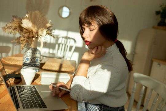 Side view of pretty young female teacher or tutor preparing for online lesson sitting at kitchen table, browsing web-pages searching for learning material, scrolling on laptop, looking at screen
