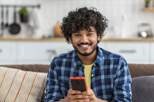 Relax at home. Young Asian man with curly hair, sitting on the couch at home, using phone to chatting online in social network, looking at the camera and smiles friendly