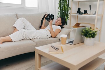 An Asian woman photographer lies on the couch with a camera and watches footage from a photo shoot at home. Freelance work