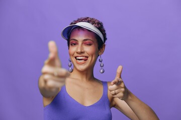 Portrait of a sporty fashion woman posing smiling with teeth and pointing a finger at the camera in a purple yoga tracksuit and a transparent cap on a purple monochrome background