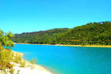 Fototapeta na wymiar Clear view of Gerosa Lake with its slightly rippled pristine glowing azure waters sitting between the greenery-topped shores and Marche hills under a cloudless blue sky on a fine summer day
