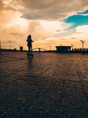 Fototapeta premium Girl is riding a push scooter on the city square against cloudy sky. Silhouette of a girl in a cityscape