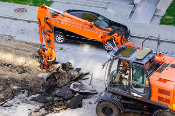 An excavator breaks up the old asphalt layer with a bucket during street repairs © Zigmunds