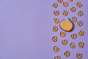 Pumpkin-shaped honey cookie and small pretzels on purple background with copy space. Festive background for Halloween, Thanksgiving and autumn holidays. Top view.