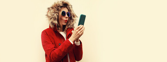 Portrait of stylish young woman with smartphone wearing red jacket with fur hood, backpack on background, blank copy space for advertising text