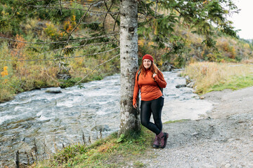 Naklejka na ściany i meble Happy redhead Woman in red hat hiking in woods. Adventure women standing near the mountain river. Freedom and active lifestyle concept. Amazing chill moment