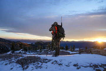 Hunter with rifle dressed in camo stands on the edge of a snowy mountain ridge looking for deer at...