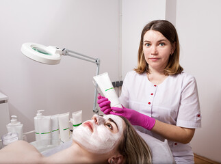 A young woman with a cosmetic white mask on her face in a beauty salon, a cosmetologist makes a care procedure with the help of cosmetics