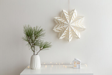 Christmas cozy winter home decor. New year interior decorations. White paper snowflake on wall, green pine branch in vase, decorative ceramic house, glowing garland lights. Composition on the dresser.