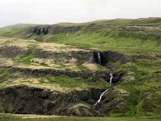 A small long waterfall with four separate sections drops down a green and beige treeless hillside in the Bifrost area of Western Iceland.