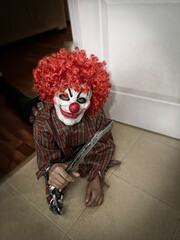 terrifying crazy clown lying on the floor with a smiling mask and a wig of curly red hair,...