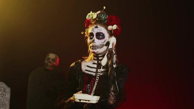 Lady of dead holding landline phone in studio, talking on office telephone with cord and wearing skull make up. Woman in santa muerte costume celebrating holy mexican ritual. Handheld shot.