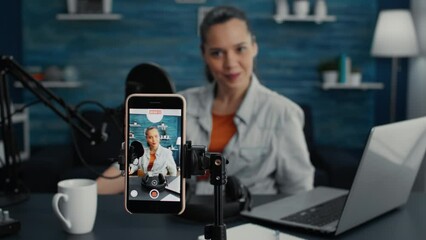 Attractive social media influencer sitting at home studio desk while starting streaming from living room with smartphone. Young content creator recording daily vlog with touchscreen modern device.