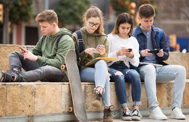 Teenage friends holding phones and browsing sitting on the stairs on the street