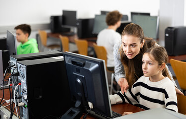 Fototapeta premium Positive female teacher helping concentrated teen schoolgirl studying in college library computer lab..