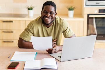 latin hispanic man looking happy while sitting with laptop and papers at kitchen