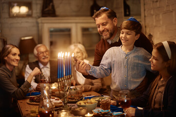 Happy father and son lighting menorah while celebrating Hanukkah with their family at dining table.
