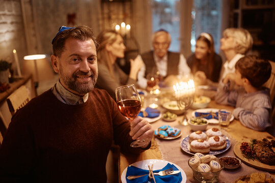 Happy Jewish Man Celebrating Hanukkah With His Extended Family At Dining Table And Looking At Camera.