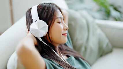 Young asian woman listening to music on couch in living room at home. Happy asia female using mobile smartphone, wearing headset and sitting on sofa