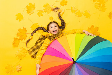 A little girl in a raincoat with a scarf lies on autumn, yellow, maple leaves and holds onto a colorful colorful umbrella. A funny child with pigtails is enjoying the autumn season.