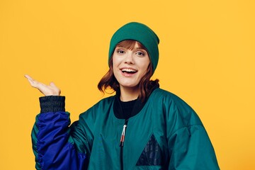 a laughing, happy woman in a green jacket and a hat in the style of the 90s stands on a yellow background, smiling happily and pointing to an empty space for inserting an advertising layout