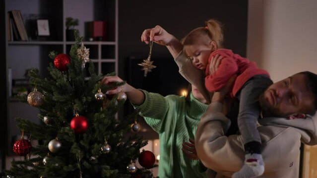 Playful hyperactive Caucasian daughter decorating Christmas tree with parents in living room. Side view portrait of cute girl enjoying leisure with young father and mother on New Year's eve